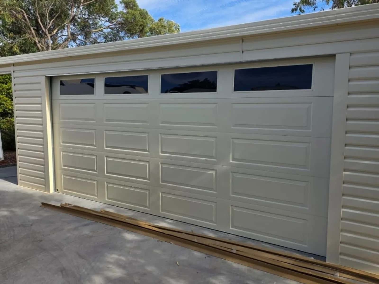 Cream panel-lift garage door with raised pressings and four tinted window inserts on a steel-clad double garage