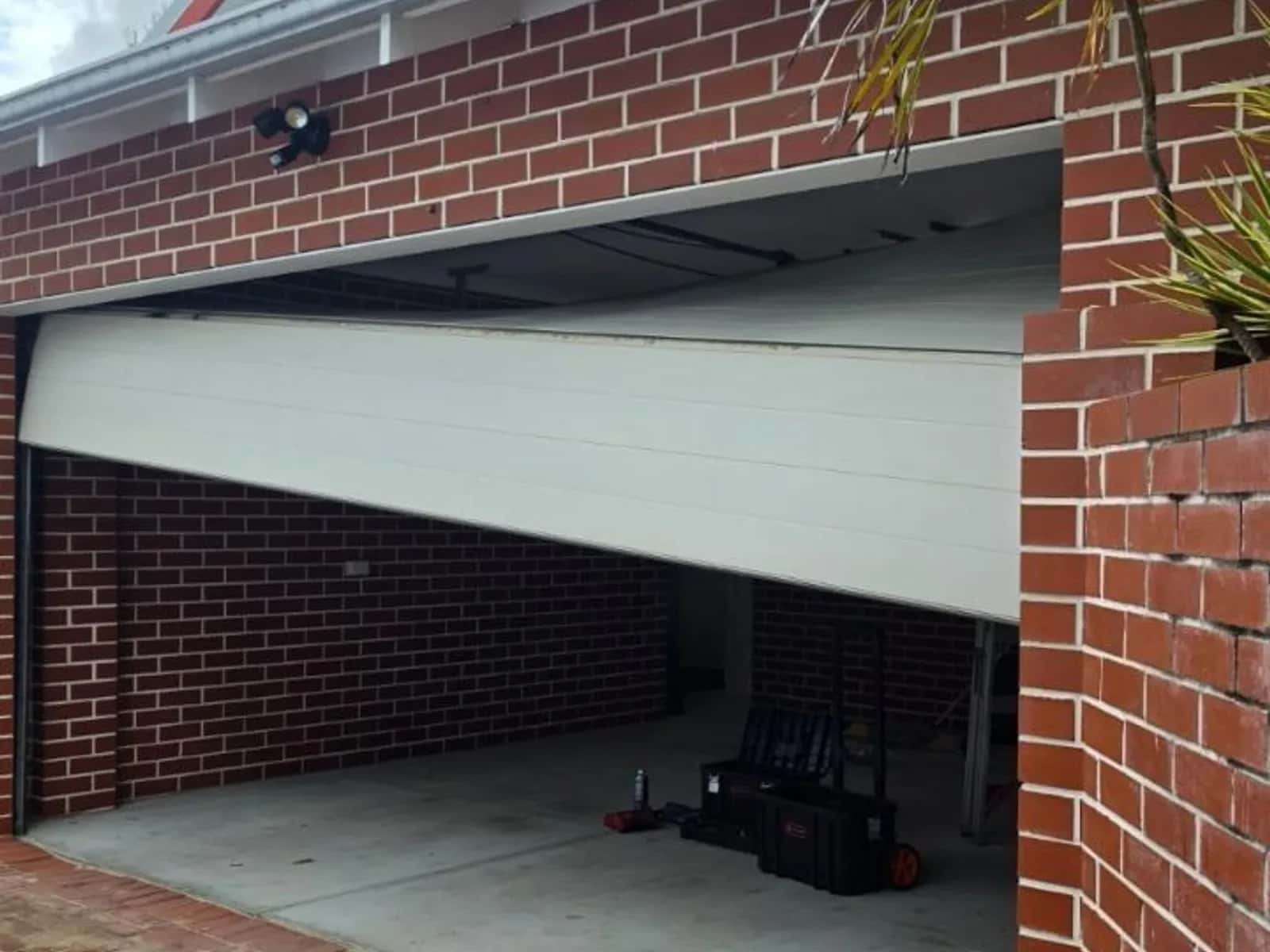 Cream one-piece tilt garage door part-open and hanging out of level on a red brick double garage
